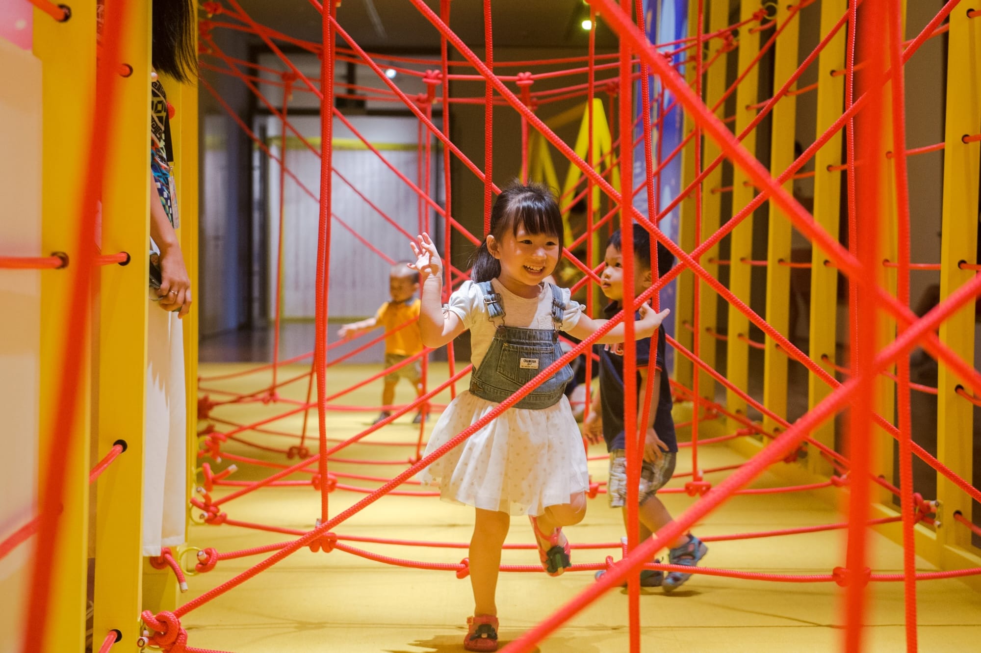 Child in OshKosh overalls plays in a maze of red ropes on yellow floor with other children.